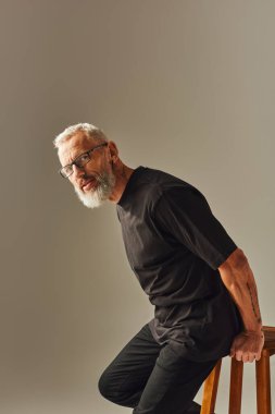 Dövmeli, gözlüklü, uzun sandalyede poz veren ve kameraya bakan olgun bir adam.