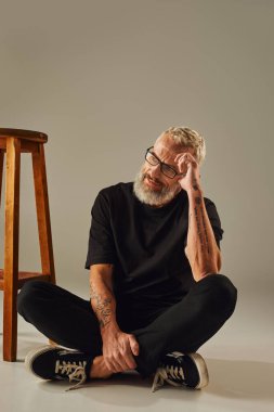 joyous handsome mature man in black t shirt with tattoos posing next to tall chair on beige backdrop clipart