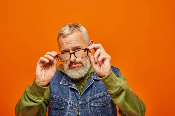 stock image handsome mature man in green vibrant hoodie and denim vest holding glasses and looking at camera