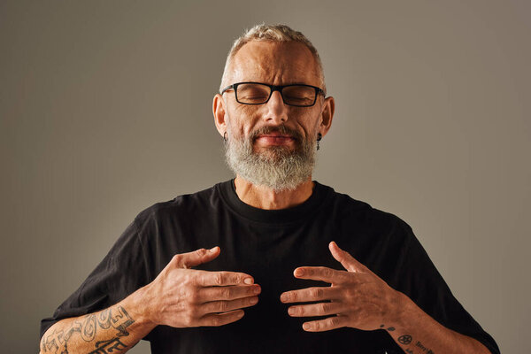 красивый веселый взрослый мужчина в черной футболке с бородой позирует с закрытыми глазами на бежевом фоне