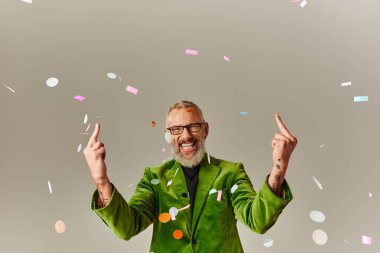 joyous mature man in green vibrant blazer with glasses showing middle fingers and smiling at camera clipart