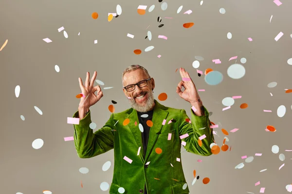 Stock image handsome jolly mature man in green blazer showing okay sign under confetti rain on beige backdrop