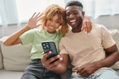 focus on smartphone, happy african american couple video chatting and waving in living room clipart