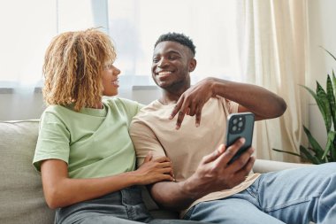 Mutlu Afro-Amerikalı adam elinde akıllı telefon tutarken kız arkadaşına işaret dili hareketi yapıyor.