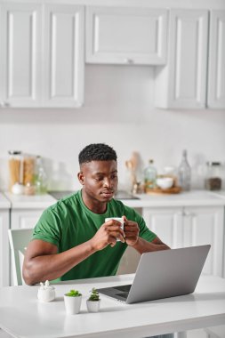 african american man enjoying cup of coffee while using laptop in kitchen, freelancer at home clipart