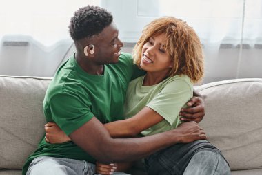 Kanepede kucaklaşan mutlu Afrikalı Amerikalı çift, işitme cihazlı sağır siyah adam ve diş teli takan kadın.