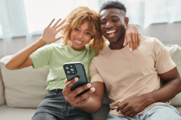 Stock image focus on smartphone, happy african american couple video chatting and waving in living room