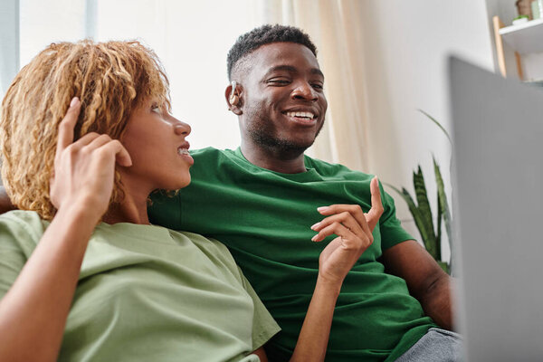 cheerful african american woman in braces talking to deaf man with hearing aid device, laptop
