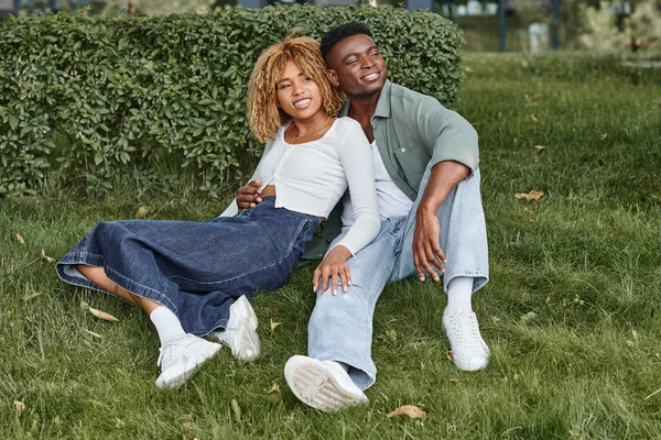 stock image cheerful african american couple hugging and sitting together on green grass, happiness