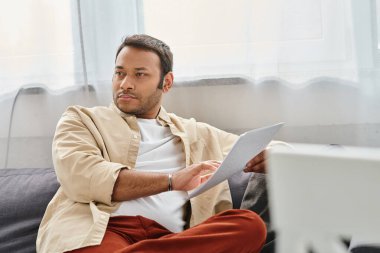 handsome indian man with blindness in cozy clothing sitting on sofa and reading braille code clipart