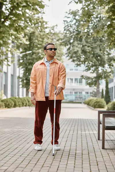 handsome indian blind man with glasses and walking stick in vivid orange jacket walking in park