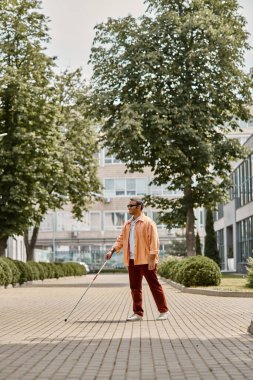 Turuncu ceketli, gözlüklü ve bastonlu neşeli Hintli kör adam parkta yürüyor.