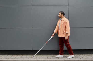 attractive indian blind man in orange jacket with helping stick walking with gray wall on backdrop clipart