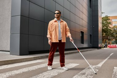 handsome indian blind man in orange jacket with glasses and stick with gray wall on backdrop clipart