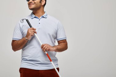 joyful indian blind man in blue tee shirt with glasses and walking stick posing on gray backdrop clipart