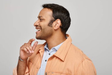 joyful indian man in vibrant orange jacket smiling happily and looking away on gray backdrop clipart