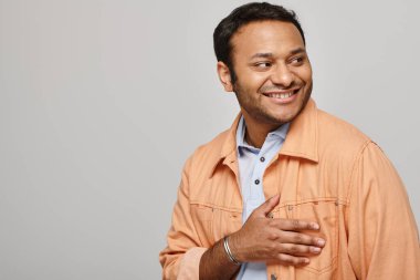 Canlı turuncu ceketli neşeli Hintli adam mutlu bir şekilde gülümsüyor ve gri arka plana bakıyor.
