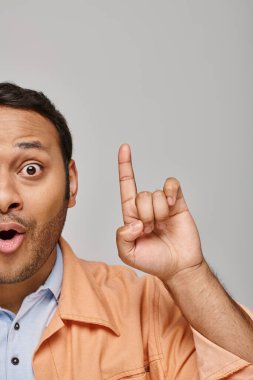 surprised handsome indian man in orange jacket posing with raised finger near face on gray backdrop clipart