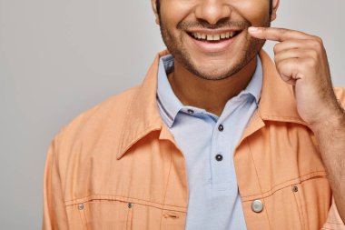cropped view of joyful indian man in orange jacket posing with finger near face on gray backdrop clipart