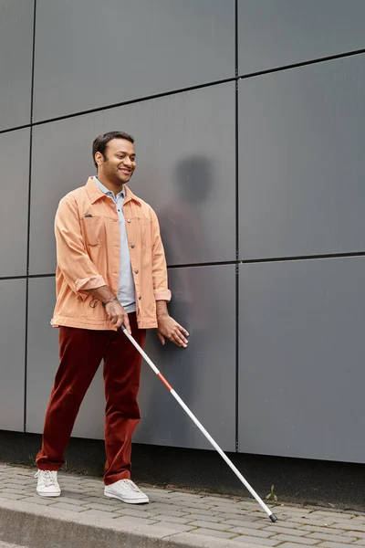 joyful indian blind man in orange jacket with helping stick walking with gray wall on backdrop