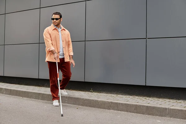 jolly indian blind man in orange jacket with walking stick and glasses with gray wall on backdrop