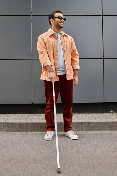 cheerful indian blind man in orange jacket with walking stick and glasses with gray wall on backdrop