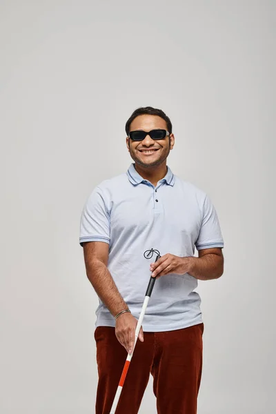 cheerful indian blind man in blue tee shirt with glasses and walking stick posing on gray backdrop