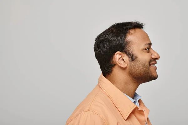 stock image merry indian man in vibrant orange jacket smiling happily and looking away on gray backdrop