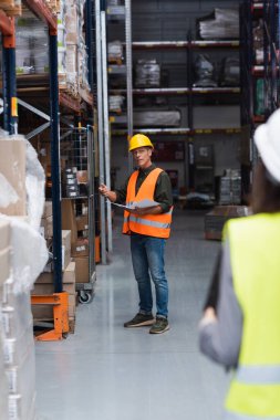 middle aged supervisor in hard hat with paperwork instructing his female employee in warehouse clipart