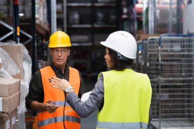 middle aged supervisor in hard hat having conversation with his female employee in warehouse clipart