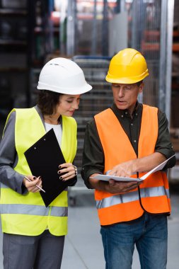 middle aged supervisor in safety vest showing paperwork to his female employee in warehouse clipart