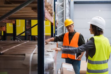 middle aged supervisor in hard hat holding tablet while explaining work to employee in warehouse clipart