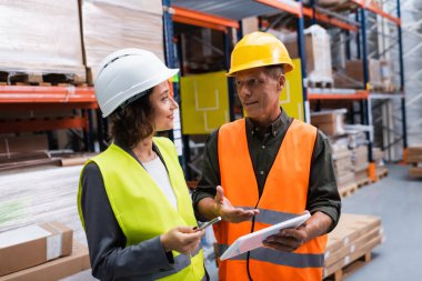 supervisor in hard hat holding tablet while discussing work to happy female employee in warehouse clipart