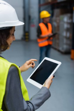Sert şapka ve güvenlik yeleği giymiş kadın depo yöneticisi elinde boş ekranlı dijital tablet tutuyor.