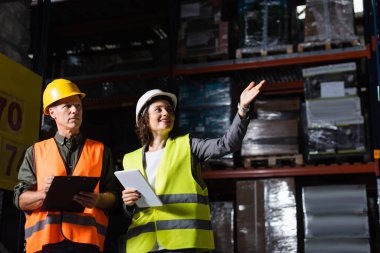 happy warehouse employee with tablet pointing away near supervisor in hard hat and safety vest clipart