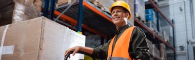 happy middle aged warehouse worker in safety vest transporting pallet with hand truck, banner clipart