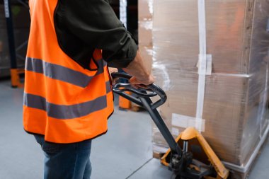 cropped middle aged warehouse worker in hard hat and safety vest transporting pallet with hand truck clipart