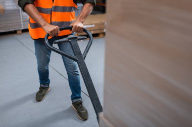 cropped photo of warehouse worker in hard hat and safety vest transporting pallet with hand truck clipart