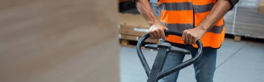cropped banner of warehouse worker in hard hat and safety vest transporting pallet with hand truck clipart