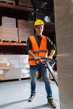 serious middle aged warehouse worker in hard hat transporting pallet with a hand truck clipart