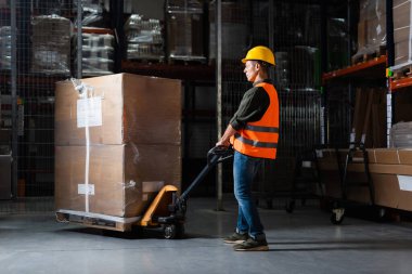strong middle aged warehouse worker in hard hat and safety vest transporting pallet with hand truck clipart