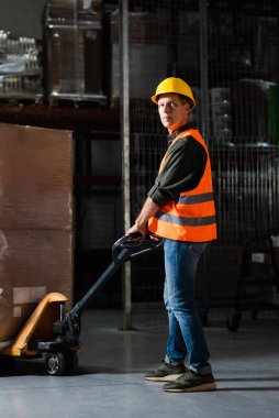 strong middle aged warehouse worker in safety vest transporting heavy pallet with hand truck clipart