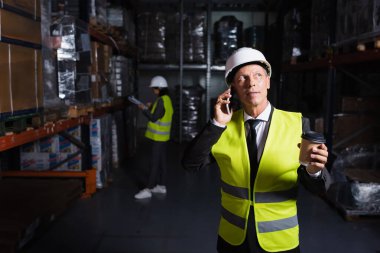 Orta yaşlı, kasketli ve güvenlik yelekli bir işçi telefon görüşmesi yaparken elinde kahve tutuyor.