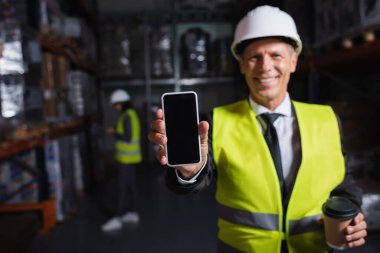 Kasketli ve güvenlik yeleği giymiş orta yaşlı mutlu bir işçi elinde kahve ve akıllı telefon tutuyor.