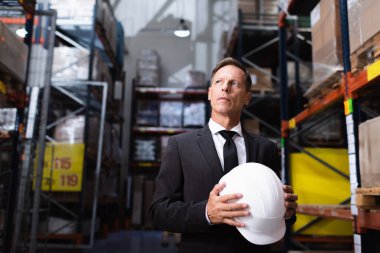 pensive middle aged businessman in suit holding hard hat in warehouse, professional headshot clipart