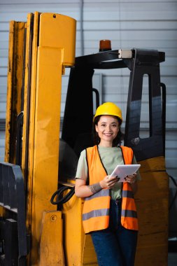 Kasketli ve çelik yelekli mutlu kadın depo çalışanı forklift ve kargo yakınında tablet tutuyor.