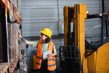 Dijital tablet tutan ve kargoyu kontrol eden çelik yelek giyen kadın depo çalışanı.