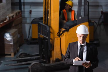 middle aged warehouse supervisor in suit and hard hat using tablet while planning logistics clipart
