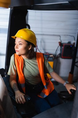 Düşünceli kadın forklift operatörü. Kasketli ve güvenlik yeleği takmış. Depoya bakıyor.