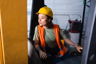Sert şapka ve güvenlik yeleği giymiş düşünceli bir kadın forklift operatörü depoya bakıyor.